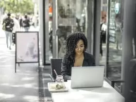 High angle of pensive African American female freelancer in glasses and casual clothes focusing on screen and interacting with netbook while sitting at table with glass of yummy drink on cafe terrace in sunny day